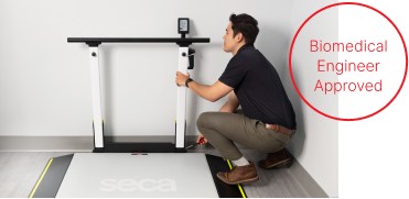 Un ingeniero biomédico se arrodilla junto a una gran plataforma de pesaje digital con bordes de seguridad y rieles de soporte verticales (seca XLine Hold). El ingeniero está realizando tareas de mantenimiento o ajustes en el equipo, con una pequeña caja de herramientas visible cerca. En la parte superior de los rieles de soporte hay una pantalla digital. A la derecha de la imagen, un círculo rojo contiene el texto «Biomedical Engineer Approved».