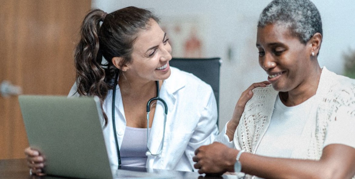 Smiling healthcare professional with stethoscope comforting a patient during a medical consultation, both sitting at a desk with a laptop.