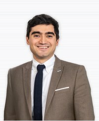 Un hombre de cabello oscuro, que lleva una chaqueta de traje marrón claro, camisa blanca y corbata oscura, sonríe mientras mira directamente a la cámara. Tiene una pequeña insignia en la solapa, y el fondo es de color neutro, con líneas horizontales sutiles.