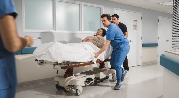 Dos profesionales sanitarios, uno con bata azul y el otro con ropa oscura, trasladan a un paciente en una camilla de hospital por un pasillo muy iluminado. El paciente, cubierto con una manta blanca, yace tranquilamente en la camilla. Un tercer profesional médico aparece en primer plano, parcialmente fuera del encuadre, sosteniendo un portapapeles. El pasillo del hospital es limpio y moderno, con ventanas y paredes blancas.