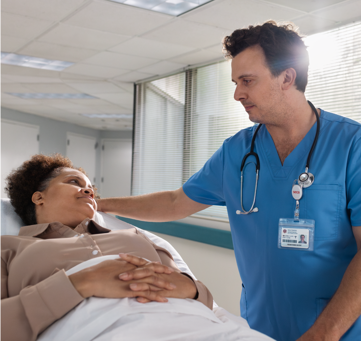 A healthcare professional in blue scrubs with a stethoscope gently places a hand on the shoulder of a patient lying in a hospital bed, offering comfort. The patient, a woman with short, curly hair, looks up at the healthcare worker, appearing calm and reassured. They are in a brightly lit hospital room with large windows in the background.
