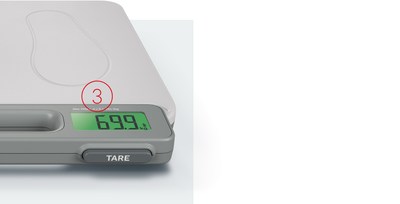 Una vista de cerca de la esquina de una báscula digital seca (seca 874+), con un círculo rojo y el número '3' resaltando la pantalla LCD que brilla en la oscuridad. La pantalla muestra un peso de '69,9 kg'. Debajo de la pantalla está el botón 'TARE', y el asa incorporada es visible a la izquierda de la pantalla. La superficie de la báscula presenta la impresión de una huella.