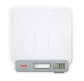 Una vista desde arriba de una báscula digital seca (seca 874+) con una superficie blanca, que presenta impresiones de dos pies. La báscula tiene un asa incorporada y una pantalla digital en la esquina inferior derecha, que actualmente muestra '0,0.' El nombre de la marca 'seca' es visible en rojo en el lado izquierdo del asa.