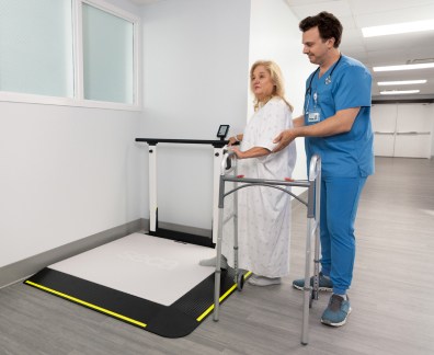 A nurse in blue scrubs assists a female patient standing on a large digital weighing platform (seca XLine Hold) with yellow safety edges. The patient, wearing a hospital gown and holding a walker, stands on the scale with the nurse providing support from behind. The scene is set in a well-lit hospital hallway near a window, with medical equipment visible in the background.