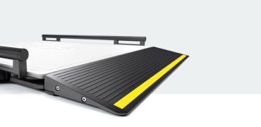 Primer plano de la rampa de una plataforma de pesaje médica de gran tamaño, con una superficie negra texturada y una franja de seguridad amarilla prominente a lo largo del borde (seca XLine Base). La rampa está ligeramente elevada y diseñada para facilitar el acceso a la plataforma de la báscula, que se ve parcialmente al fondo.