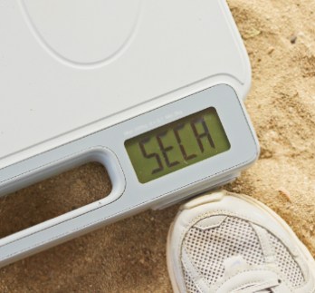 Una toma de cerca de una báscula seca (seca 874+) colocada sobre suelo arenoso. La pantalla digital muestra el nombre de la marca 'seca,' y se puede ver parte de una zapatilla blanca de una persona al lado de la báscula.