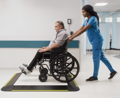 Una enfermera con bata azul empuja a un paciente varón en silla de ruedas hacia una gran plataforma de pesaje digital (seca XLine Base) con bordes de seguridad amarillos, situada en el pasillo de un hospital. El paciente está cómodamente sentado en la silla de ruedas y la enfermera le guía atentamente hasta la báscula. La escena tiene lugar en un centro médico limpio y moderno.
